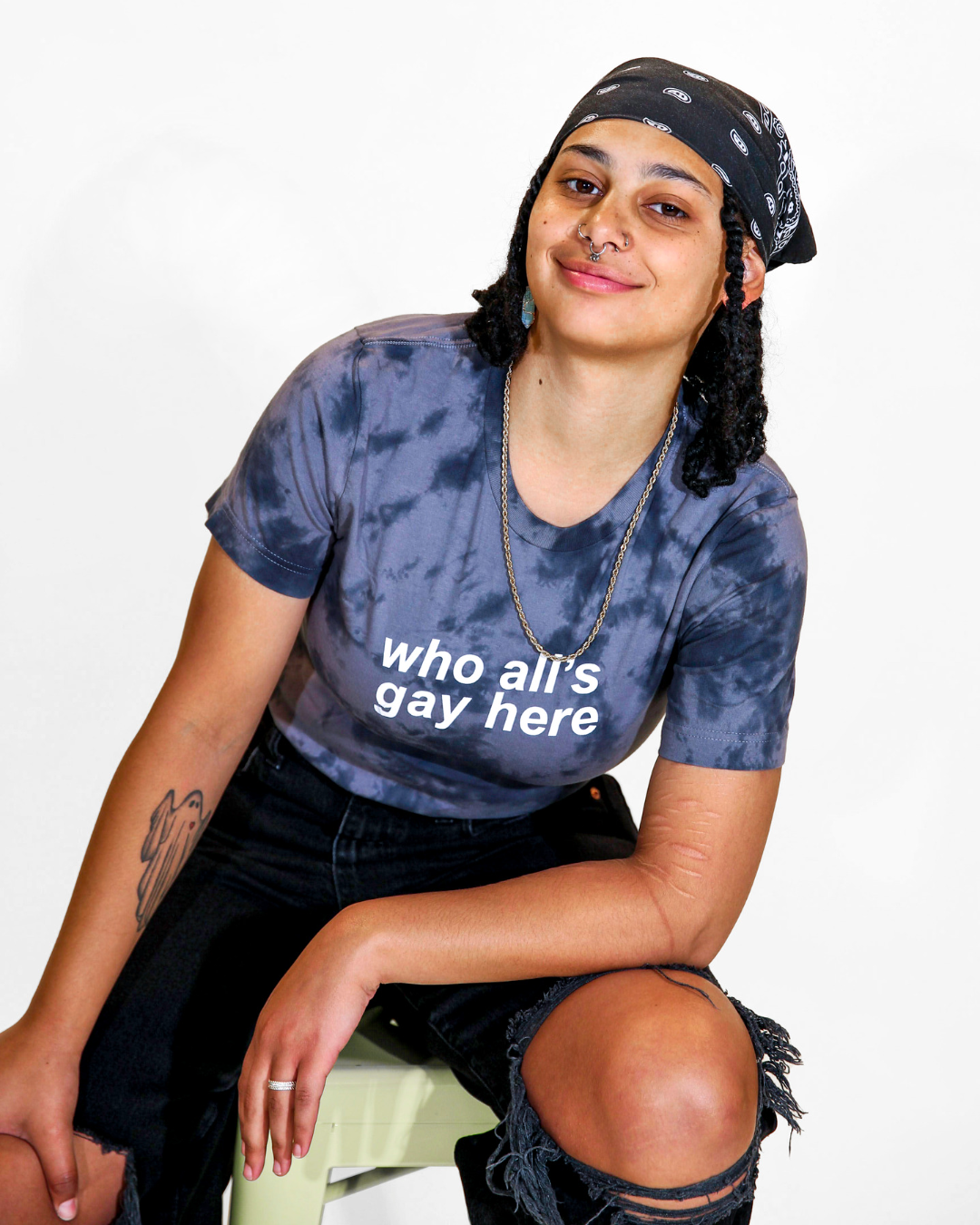 Model Grey is wearing the Who All's Gay Here Tie-Dye Tee in size S. They are 5'4" and their bra size is 32D. The tee is black and grey in a tie-dyed pattern. There is white sans-serif text that says "Who All's Gay Here." The text is askew at a diagonal in two lines. The design is by Erin Sullivan.
