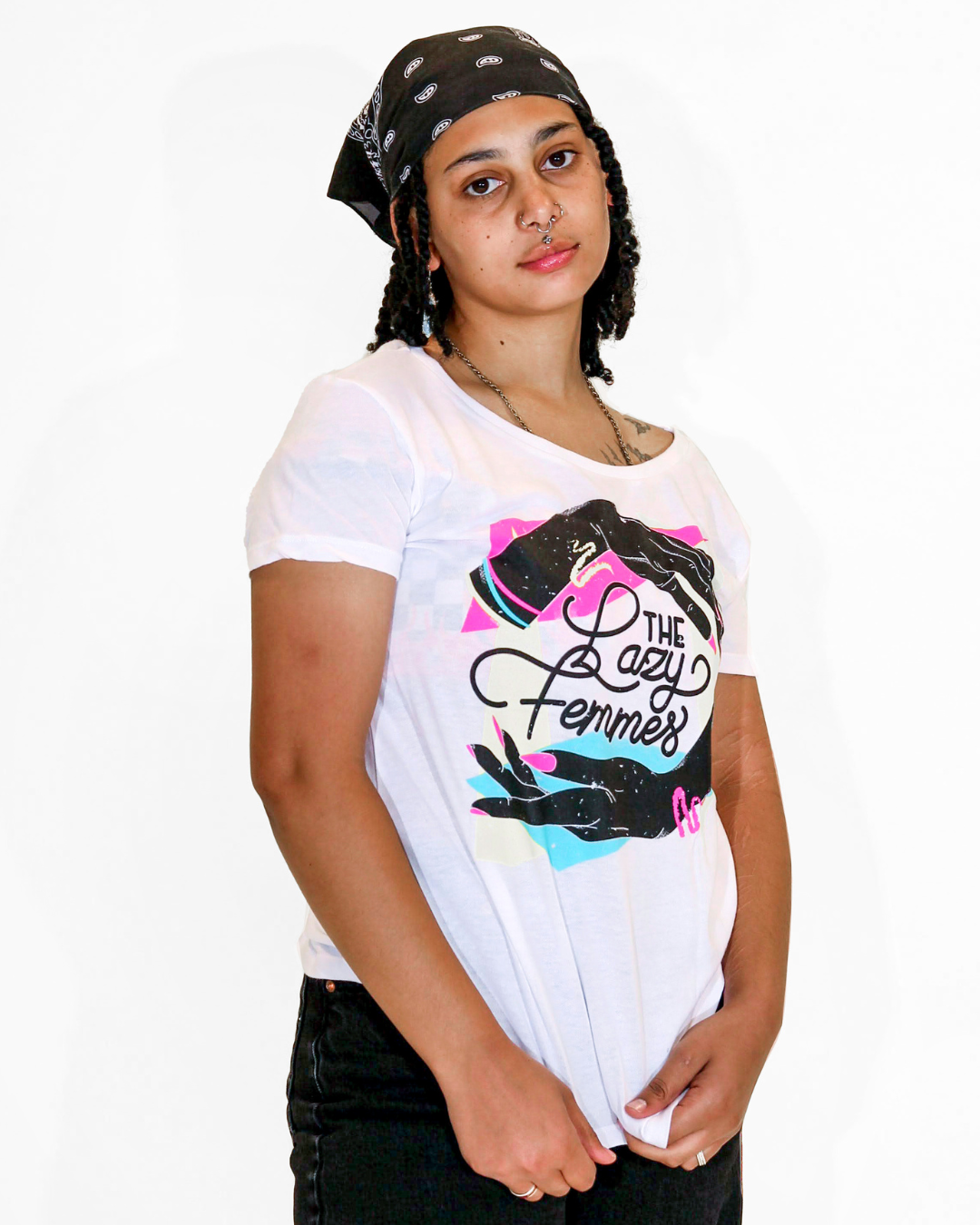 Model Grey is wearing The Lazy Femmes Band Tee in size S. They are 5'4" and their bra size is 32D. The tee is white with a multi-color graphic of two hands encircling the words "The Lazy Femmes" in black loopy cursive. The hands have neon pink, yellow, and blue accessories like nails, rings, and bracelets.