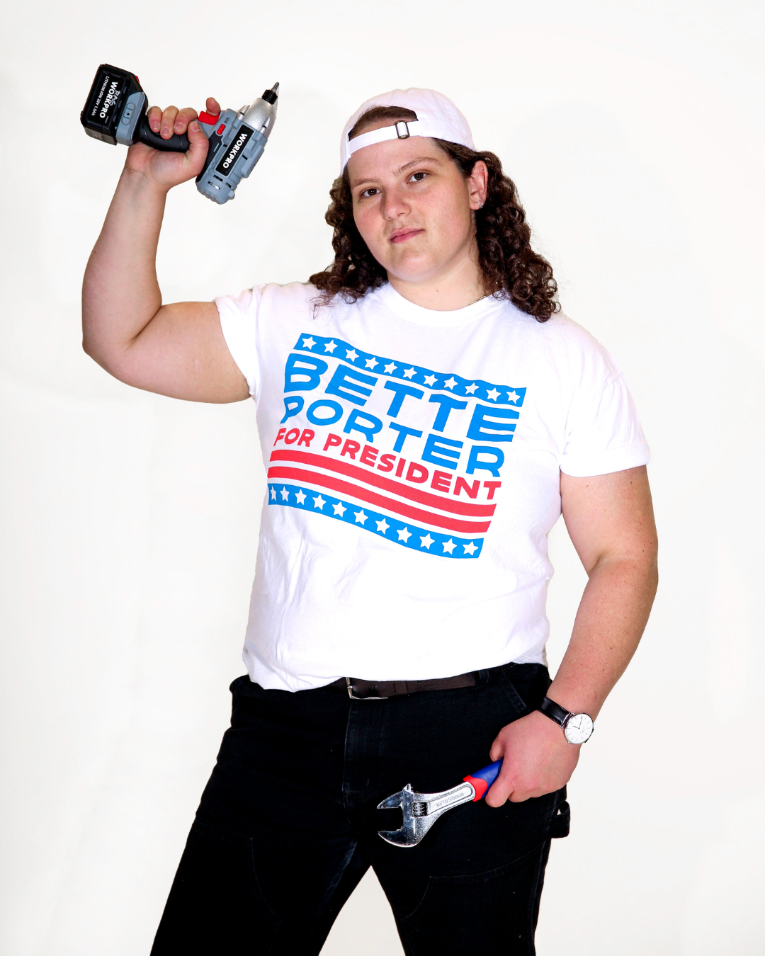Model Jenny is wearing the Bette For President Tee in size XL. They are 5'9" and their bra size is 38DDD. They are also holding a power drill on one hand and a wrench in the other. The tee is white and has a blue and red graphic that resembles a political campaign sign. In the middle are the words "Bette Porter For President," referring to the character Bette Porter from the tv show "The L Word" on Showtime.