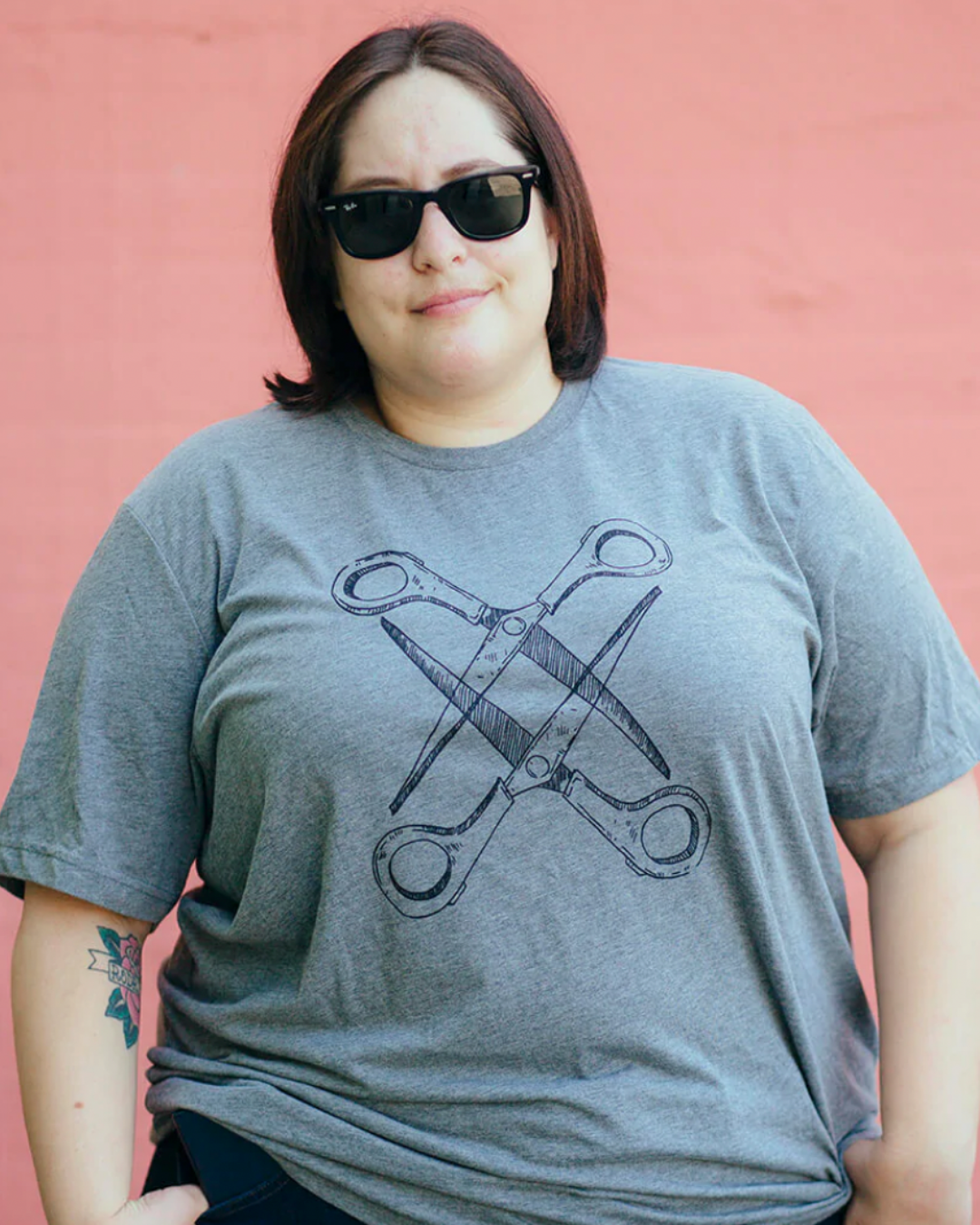 Model Justine is wearing the Grey Scissoring Tee in size 3XL. She is 5'5" and her bra size is 42DD. The tee is grey with a graphic of two scissors in a vertical alignment. Their blades are open and overlapping each other. The graphic is outlined in dark grey.