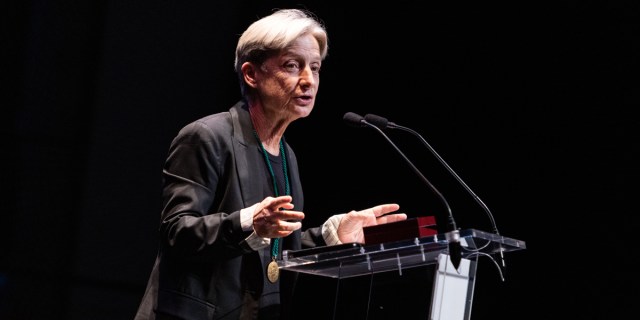 Judith Butler talks at a podium in a black suit against a black background