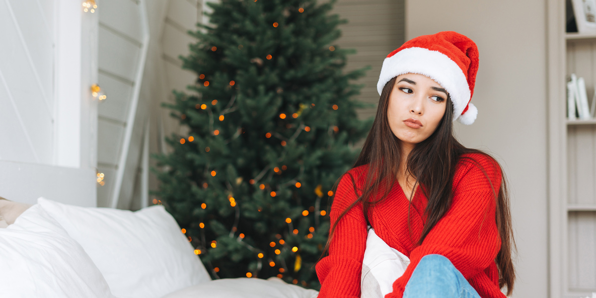 A young Asian woman with long hair and a Santa hat makes a very sad face while sitting on a bed in front of a Christmas tree.