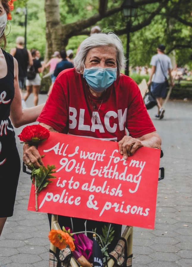 Shatzi Weisberger, known as The People's Bubbie, at another one of the 2020 Black Lives Matter uprisings. This time she wears a red t-shirt, a blue mask, and holds a red rose and a red sign with white lettering that says All I Want For My 90th Birthday Is To Abolish Police And Prisons
