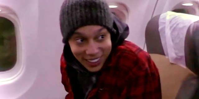 Brittney Griner is smiling, in a red coat and grey beanie, and a hoodie, she is on a plane returning back to the United States from Russia.