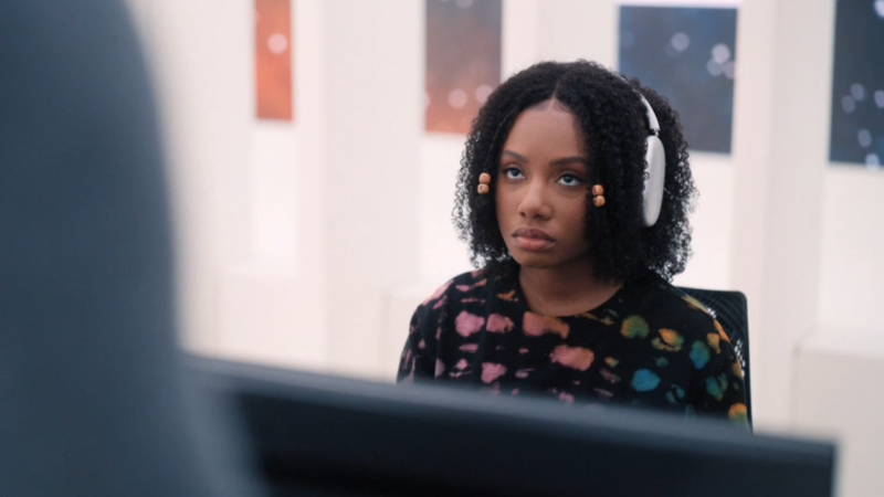 Dana sits at her desk with headphones on, unamused