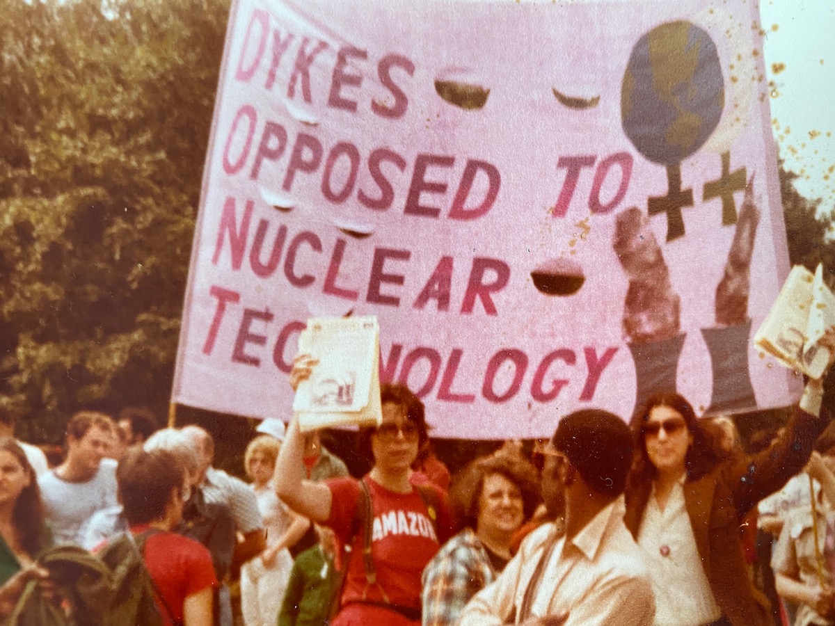 Scanned image of Shatzi at a Dykes Opposed to Nuclear Technology demonstration — many protestors fill the foreground, and a huge banner reading Dykes Opposed to Nuclear Technology with an image of a globe is held up in the background. The photo is scanned.
