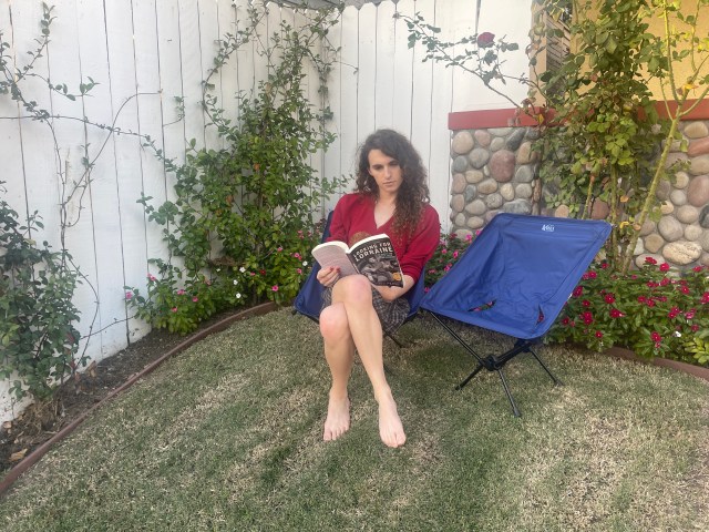 Autostraddle writer Drew Gregory in her backyard, sits in a dark blue foldable, cloth, camping chair, contentedly reading a book