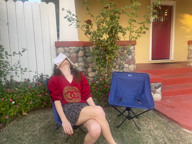 Autostraddle writer Drew Gregory in her backyard, sits in a dark blue foldable, cloth, camping chair, with a book resting on her head while she dozes