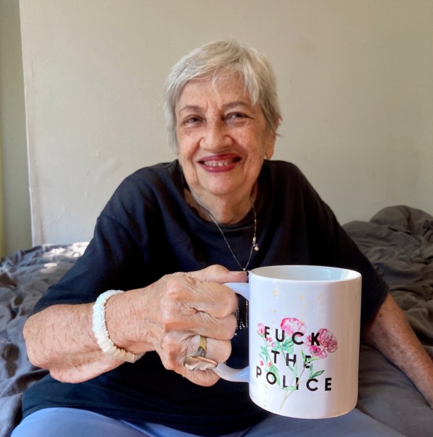 Shatzi Weisberger, known as The People's Bubbie, smiling and holding up a white mug that says Fuck The Police with beautiful flowers in the background