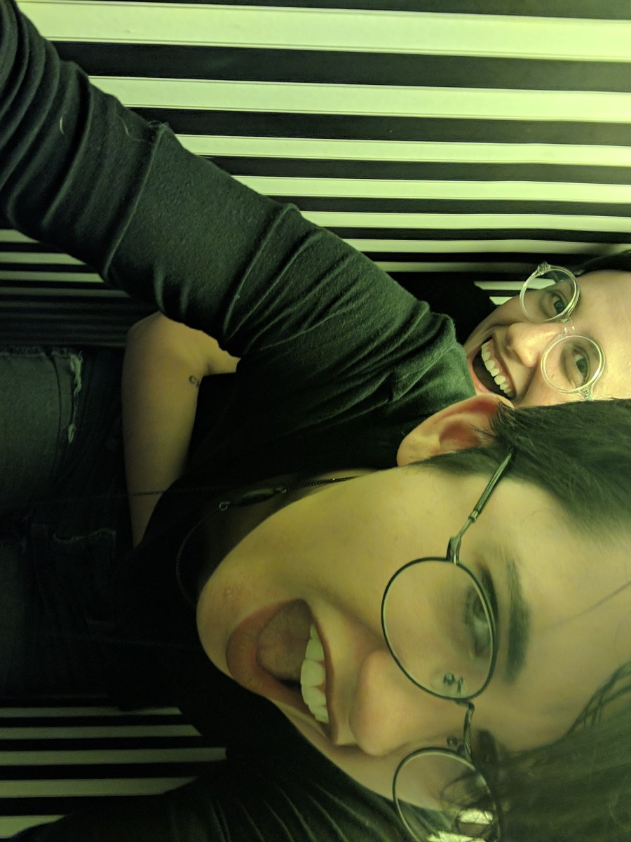 Nico and Sadie are in a greenlit striped hallway that is very narrow inside of an art house. Sadie grabs Nico from behind and Nico makes a mock affronted face.