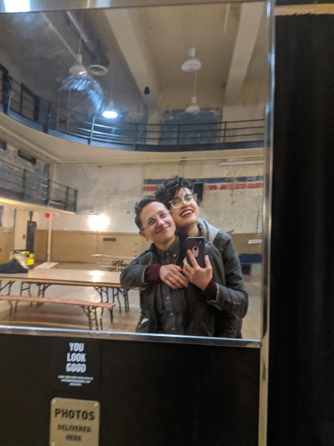 Sadie and Nico take a photo in a mirror in a gymnasium room at the ace hotel. Nico is holding Sadie from behind. They are smiling.
