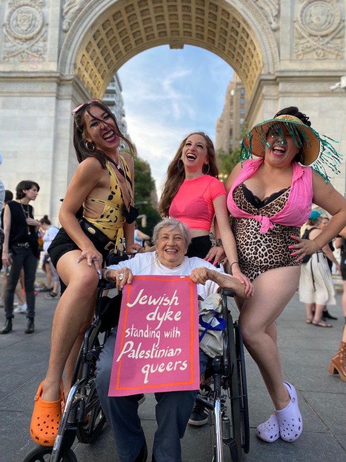Shatzi Weisberger, known as The People's Bubbie, sits in her wheelchair in Washington Square Park holding a pink sign that reads Jewish dyke standing with Palestinian queers. She's smiling, and the three young queer people posing with her are smiling and laughing, too.