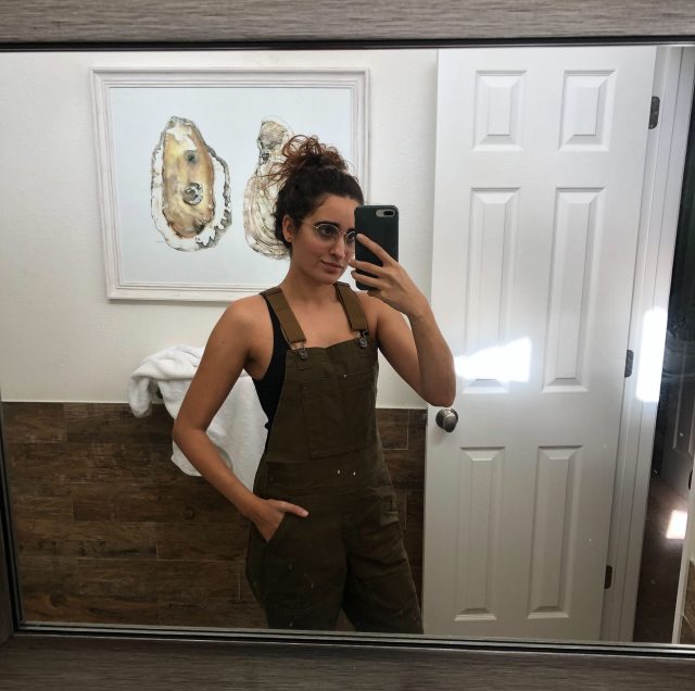 Autostraddle Kayla Upadhyaya Kumari stands in front of a mirror in her apartment, taking a selfie that shows off her new dark green overalls from REI 