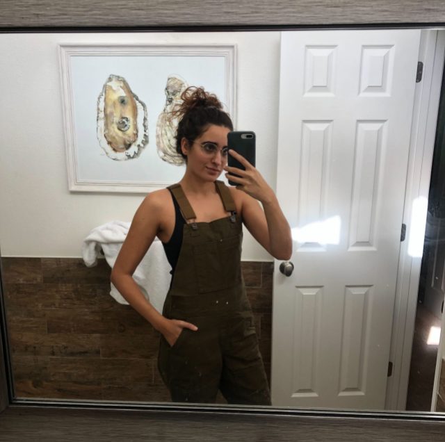 Autostraddle Kayla Upadhyaya Kumari stands in front of a mirror in her apartment, taking a selfie that shows off her new dark green overalls from REI 