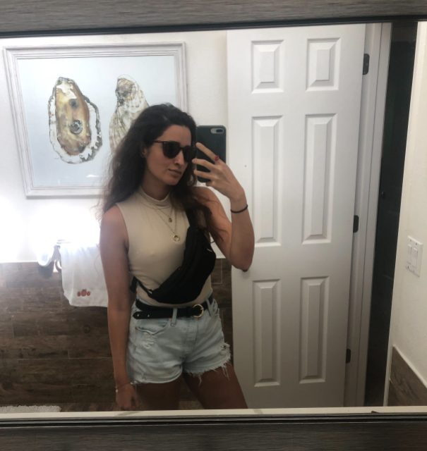 Autostraddle Kayla Upadhyaya Kumari stands in front of a mirror in her apartment, taking a selfie that shows off her new black fanny pack from REI 