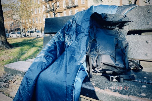A dark blue, insulated, puffy blanket from REI rests on a park bench outdoors, on a sunny, crisp, cold day in New York City