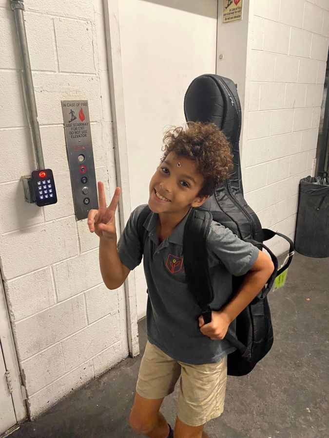 Sai's kid posing with their cello, making a peace sign with their fingers, looking cute as ever