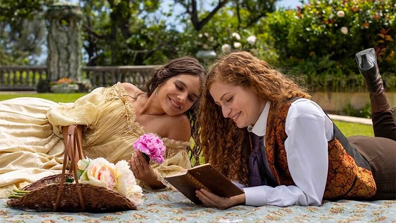 Screenshot from Fantasy island: Two women in old-timey outfits (one traditionally masculine, one traditionally feminine) lay on a blanket outside, reading together.