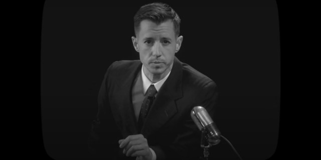 Chase Joynt as Garfinkel in Framing Agnes. He wears a suit and looks at the camera framed in a black and white TV set.