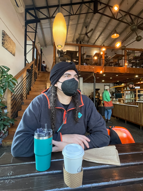 Autostraddle writer Abeni Jones sits in a cozy cafe, wearing a mask, with a Stanley mug in front of her, as well as a cup of coffee in a to-go cup