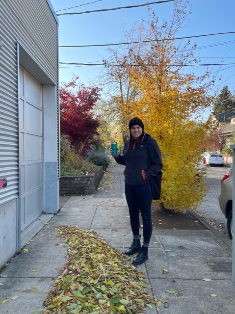 Autostraddle writer Abeni Jones stands outside on a Fall day with colorful yellow leaves, holding a Stanley coffee mug from REI and grinning