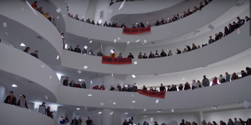 A protest at the Guggenheim. Crowds of people as prescription slips fall from above. Red banners say Shame on Sacklers and 400,000 dead.