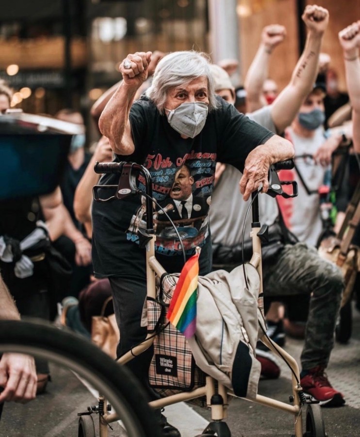 Shatzi Weisberger, known as The People's Bubbie, at one of the 2020 Black Lives Matter uprisings. She is masked, with her fist in the air, leaning against her walker. There is a rainbow flag on her walker and many other protestors are around her with their fists in the air, too.