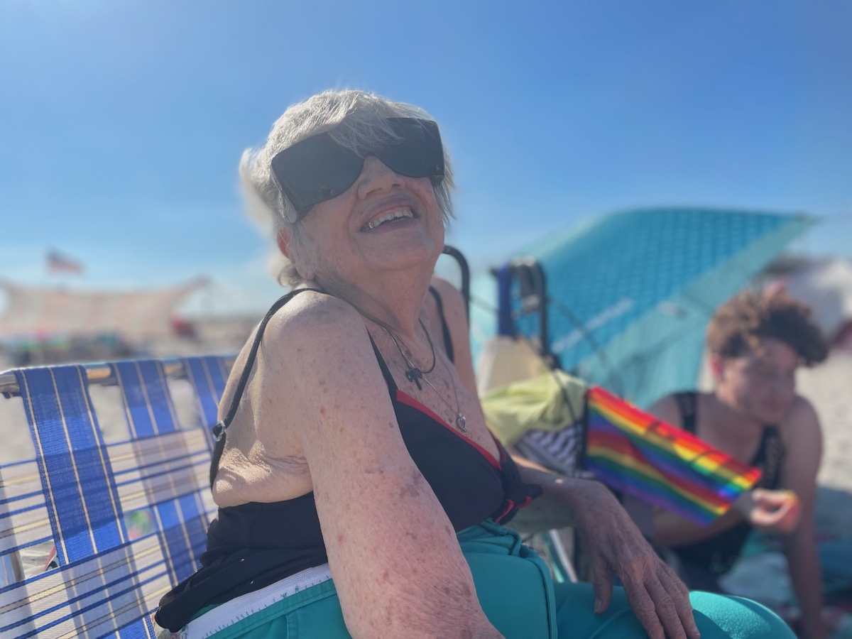 Shatzi Weisberger, known as The People's Bubbie, posing for a photo during Pride at Riis Beach. She is wearing dark sunglasses and a bathing suit and smiling at the camera as the sun lights up her whole face. A rainbow flag and some friends sit in the background.