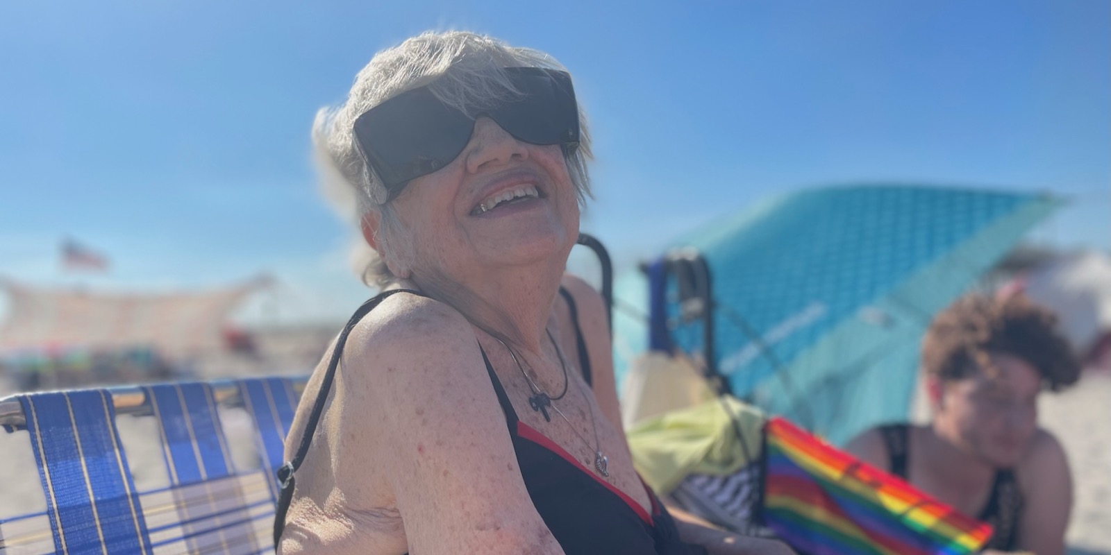 Shatzi Weisberger, known as The People's Bubbie, posing for a photo during Pride at Riis Beach. She is wearing dark sunglasses and a bathing suit and smiling at the camera as the sun lights up her whole face. A rainbow flag and some friends sit in the background.