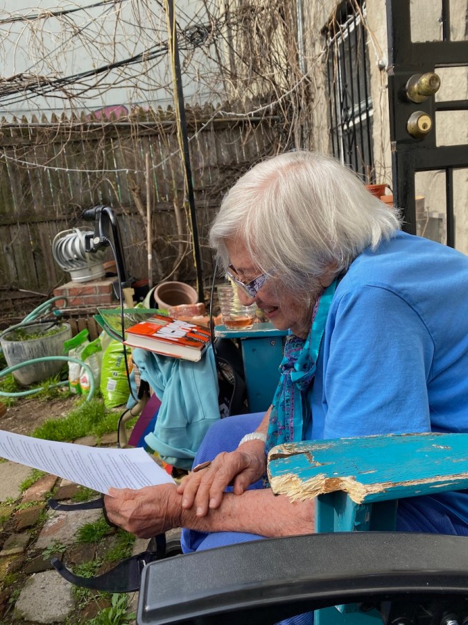 Shatzi Weisberger, known as The People's Bubbie, reading her obituary. She wears a blue t-shirt with a blue scarf around her neck, and she's smiling as she holds the paper that contains the text you are currently reading