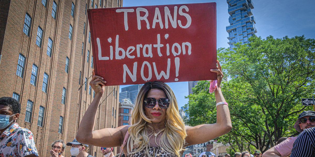 A Black trans woman in sunglasses and a leopard print tank top holds a sign that says "Trans Liberation Now!"