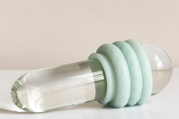 Against a beige background, a glass dildo rests on a white surface. Around the base of the dildo, there is a light blue Ohnut ring.