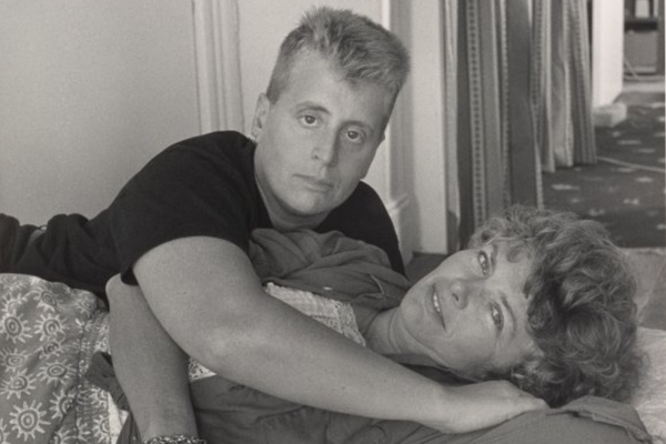 A black and white image shows Minnie Bruce Pratt, a white woman with curly short hair and bangs, lying on her back on a bed. The bed is covered in a quilt. She's wearing a dress printed with small suns and a cardigan, and she's smiling at the camera. Leslie Feinberg, a masculine-presenting white person with short hair wearing a black T-shirt, leans over Minnie and looks at the camera with a serious expression. There is a mirror and curtains in the background.