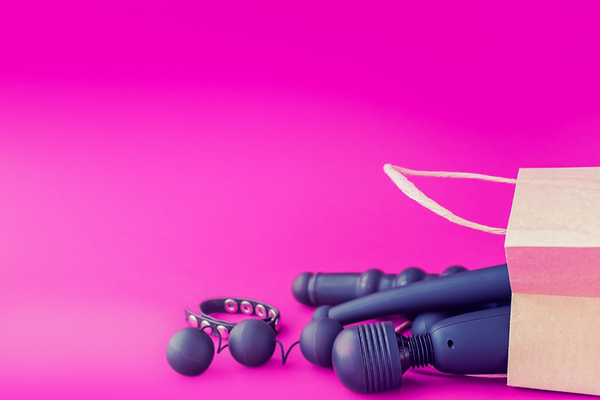 Against a hot pink background, a paper bag is turned over on its side. Purple sex toys, including an internal vibrator, a wand vibrator, Kegel balls, and a cock ring, spill out.