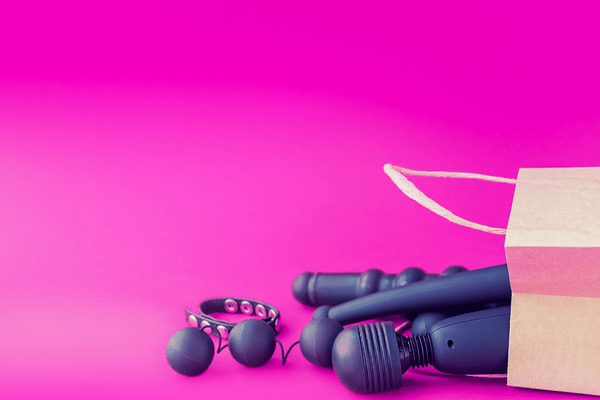 Against a hot pink background, a paper bag is turned over on its side. Purple sex toys, including an internal vibrator, a wand vibrator, Kegel balls, and a cock ring, spill out.