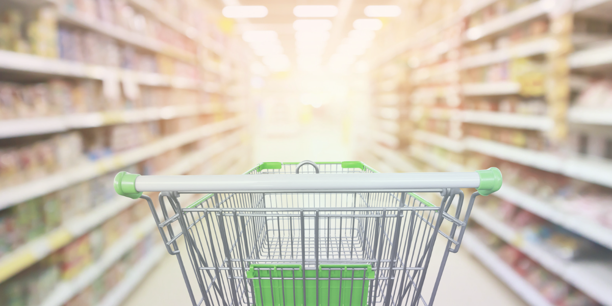 a silver and green shopping cart sits in the center of a grocery store aisle, with snacks visible on the shelves to either side