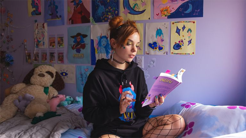 Quinni sits on her bed in front of brightly colored paintings, reading a book