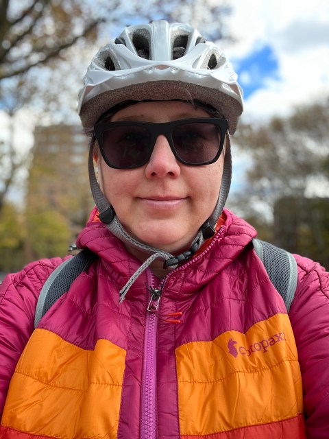 Close-up selfie of Autostraddle writer Heather Hogan smiling while wearing a grey bike helmet and a brightly colorful Cotopaxi jacket 
