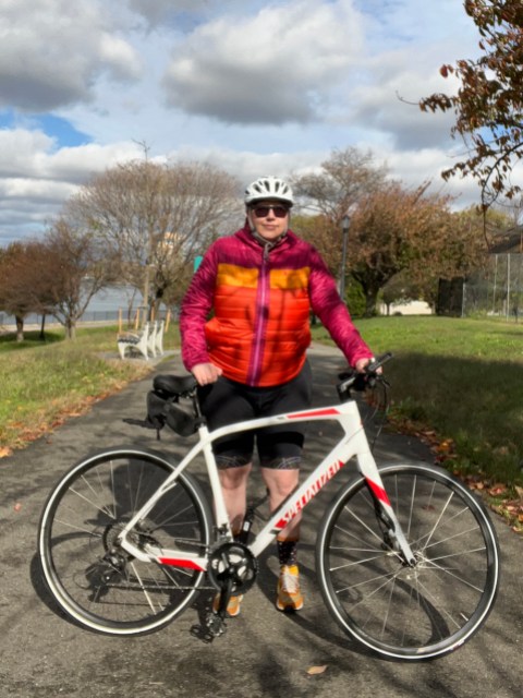 Autostraddle writer Heather Hogan stands beside her Specialized white road bike, wearing a bright orange and pink, puffy Cotopaxi jacket from REI and a grey bike helmet