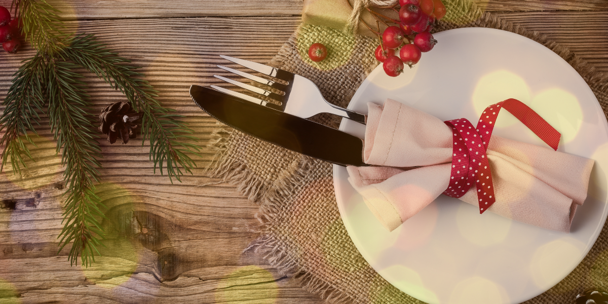 Silverware on a white plate with a red ribbon tied around them. There are cranberries and a piece of a Christmas tree on the table next to the plate.