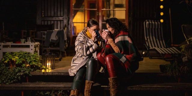 Two women huddled for warmth on the steps of a cabin, draped in blankets and sipping out of mugs