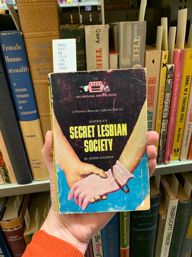 a lesbian pulp novel cover that reads Secret Lesbian Society and has a photo of two white women holding hands, one is wearing a glove with a pretty bracelet and one is not