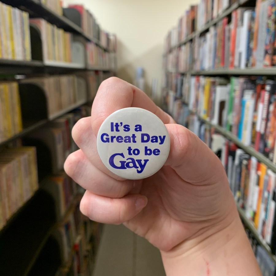 a white hand holds a button that reads "It's A Great Day To Be Gay," and we can tell they're in the archives, with many books and bookshelves behind them