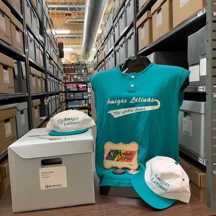 a photo of the Gerber/Hart Archives, featuring many shelves with many boxes in the background, and in the foreground materials from the Amigas Latinas collection: a bright blue tank top and a blue and white hat with the words Amigas Latinas on them, next to a closed box of other materials