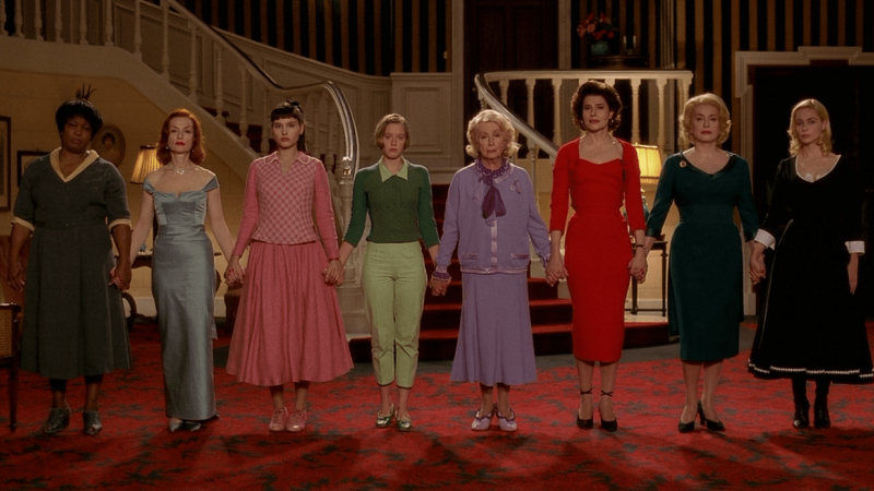 Eight women in different colored outfits hold hands in front of a staircase. 