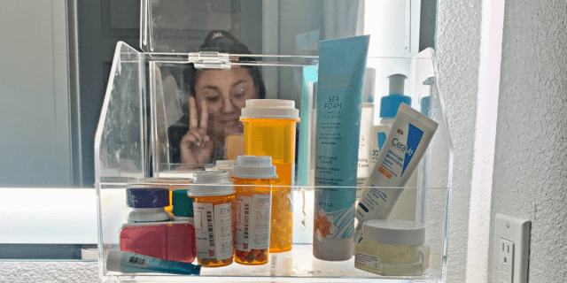 neurodiverse life hacks: a close up of clear bathroom storage containers, filled with knickknacks, stacked on top of each other. The author is making a peace sign in the back.