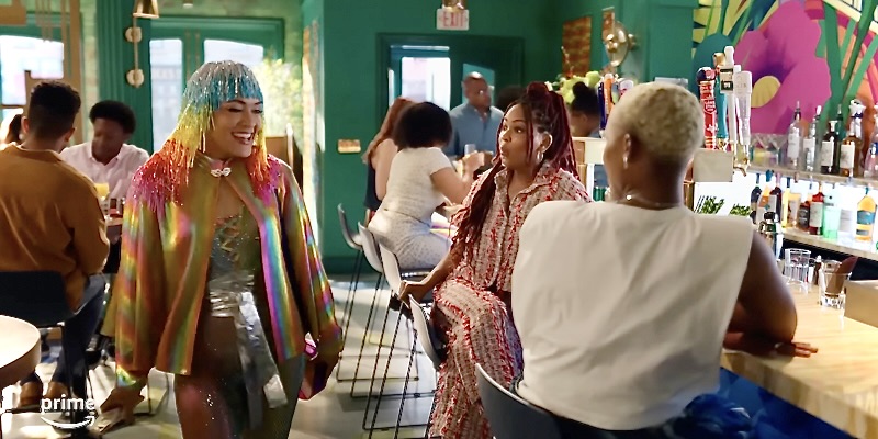 In a screenshot from Harlem Season Two, Quinn is dressed in a head to do rainbow (including a sparkly wig) for Pride, she's smiling and showing off for her friends, who are looking at her with faces of surprise, but also like her outfit is ugly and they don't have the heart to tell her.