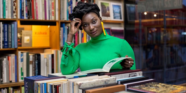 A black woman in a green sweater is in a library, reading a book.