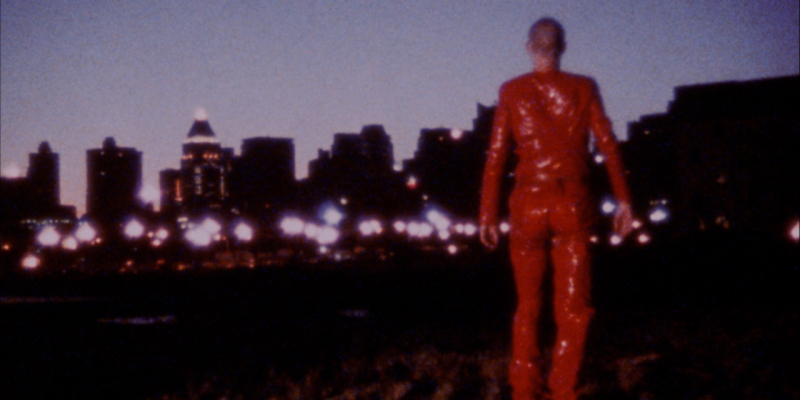 A person in red latex stands with their back to the camera looking at a dark city.