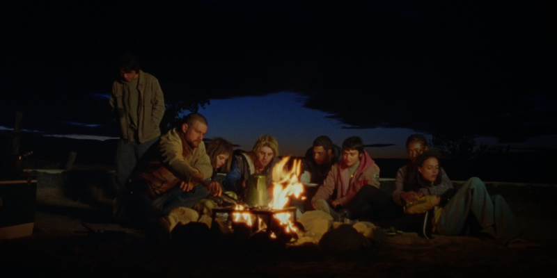 A group of young people sit around a fire in the dark.